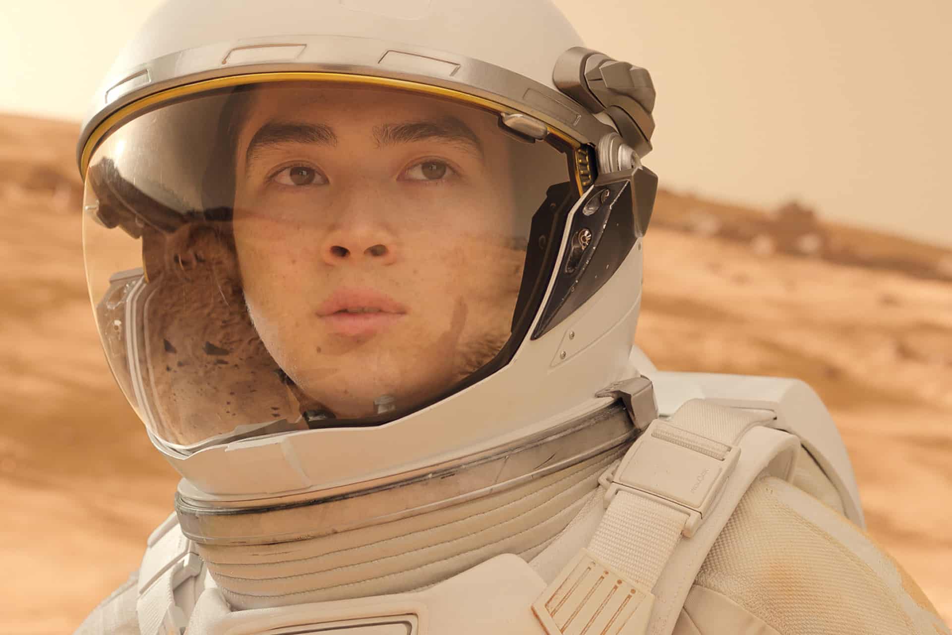 A person in a white spacesuit and helmet looks thoughtfully into the distance, standing on a dusty, reddish landscape resembling the surface of Mars—evoking scenes from For All Mankind Season 5. The background is blurred with rocky terrain.