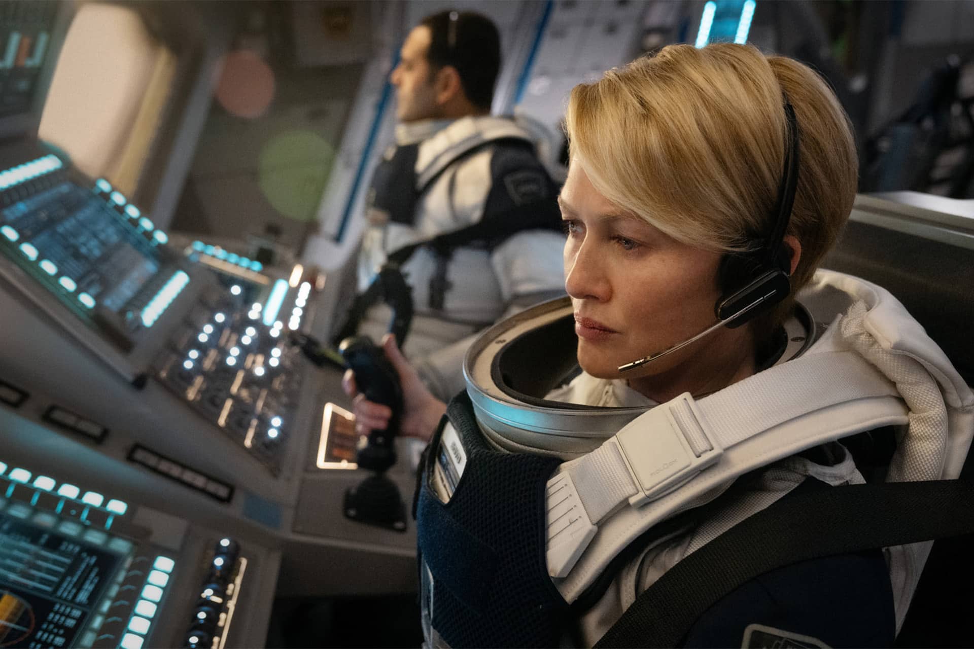 A woman in a spacesuit with a headset sits at a control panel inside a spacecraft, focused on the controls—reminiscent of For All Mankind Season 5. Another astronaut is seated beside her, both surrounded by buttons and screens.