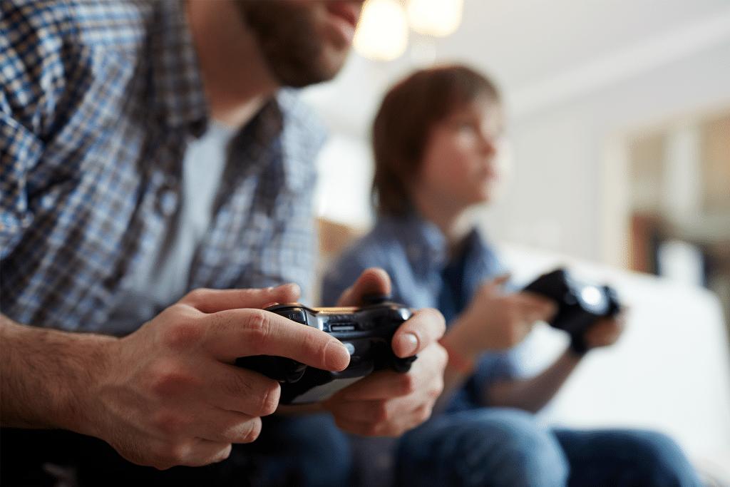 A man and a child sit side by side, holding Apple TV Console controllers and playing a game together. The focus is on their hands and controllers, with their faces out of focus in the background.