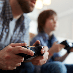 A man and a child sit side by side, holding Apple TV Console controllers and playing a game together. The focus is on their hands and controllers, with their faces out of focus in the background.