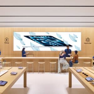 A modern electronics store with light wood decor, display tables of devices, and a large screen showing a tablet with wings. Store staff in blue shirts assist customers at counters, including the Genius Bar and "Pickup" areas.