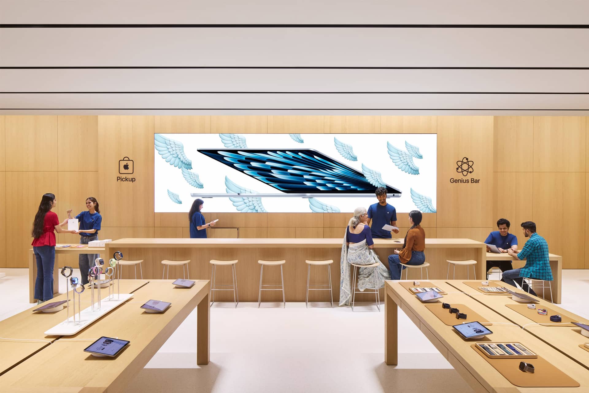 A modern electronics store with light wood decor, display tables of devices, and a large screen showing a tablet with wings. Store staff in blue shirts assist customers at counters, including the Genius Bar and "Pickup" areas.