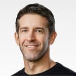 A man with short dark hair and light stubble, rumored to be Apple Next CEO, smiles at the camera. He is wearing a black crew-neck shirt and is posed in front of a plain, light background.
