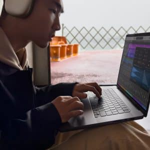 A person wearing headphones is using a laptop, editing audio tracks in Apple Creator Studio. They are seated indoors near a window, with an orange chair visible outside.