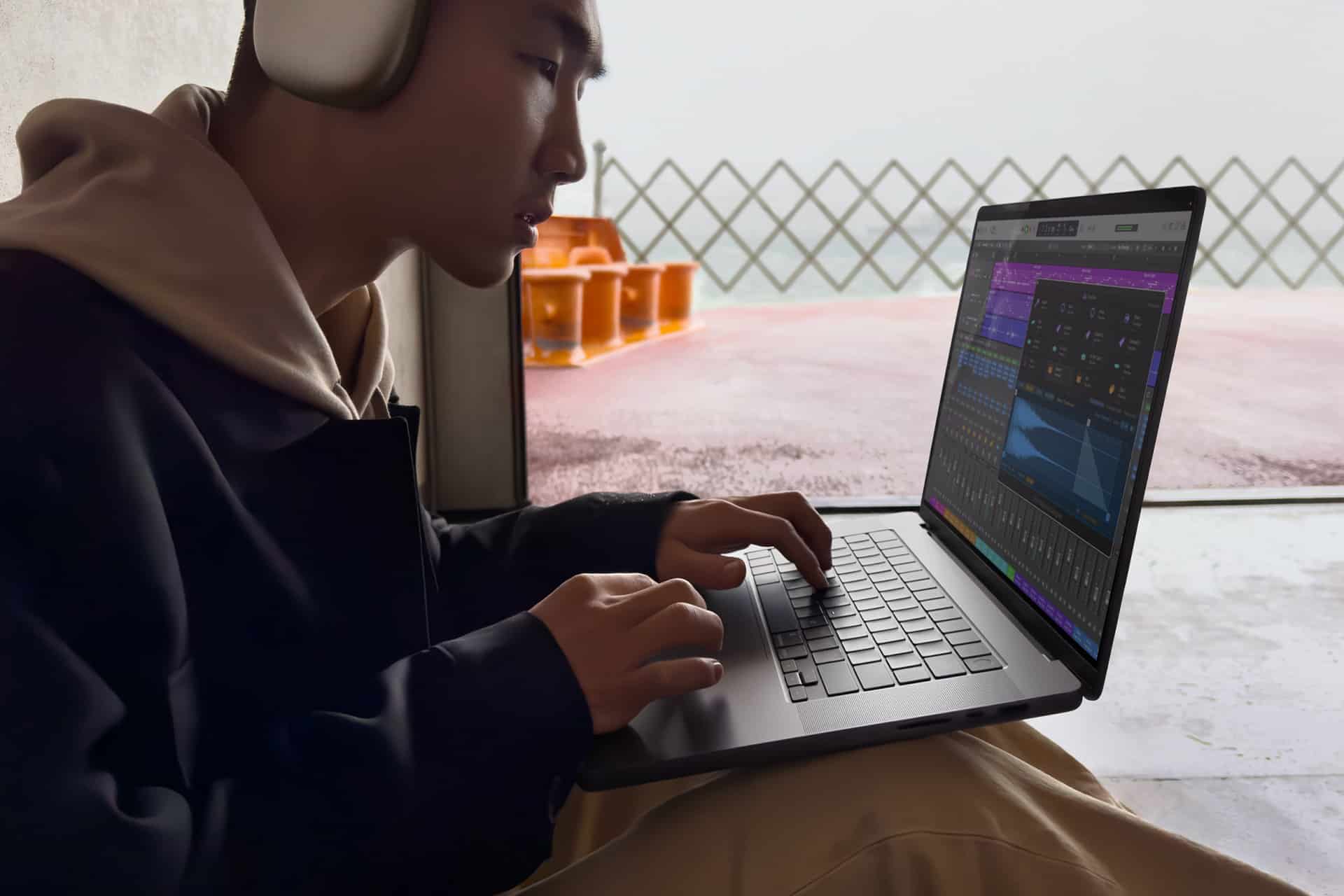 A person wearing headphones is using a laptop, editing audio tracks in Apple Creator Studio. They are seated indoors near a window, with an orange chair visible outside.