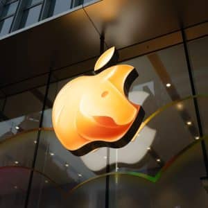 A large, illuminated Apple logo shines on the exterior glass facade of an Apple Store, with building reflections and colorful wavy lines celebrating Apple Lunar New Year along the bottom.