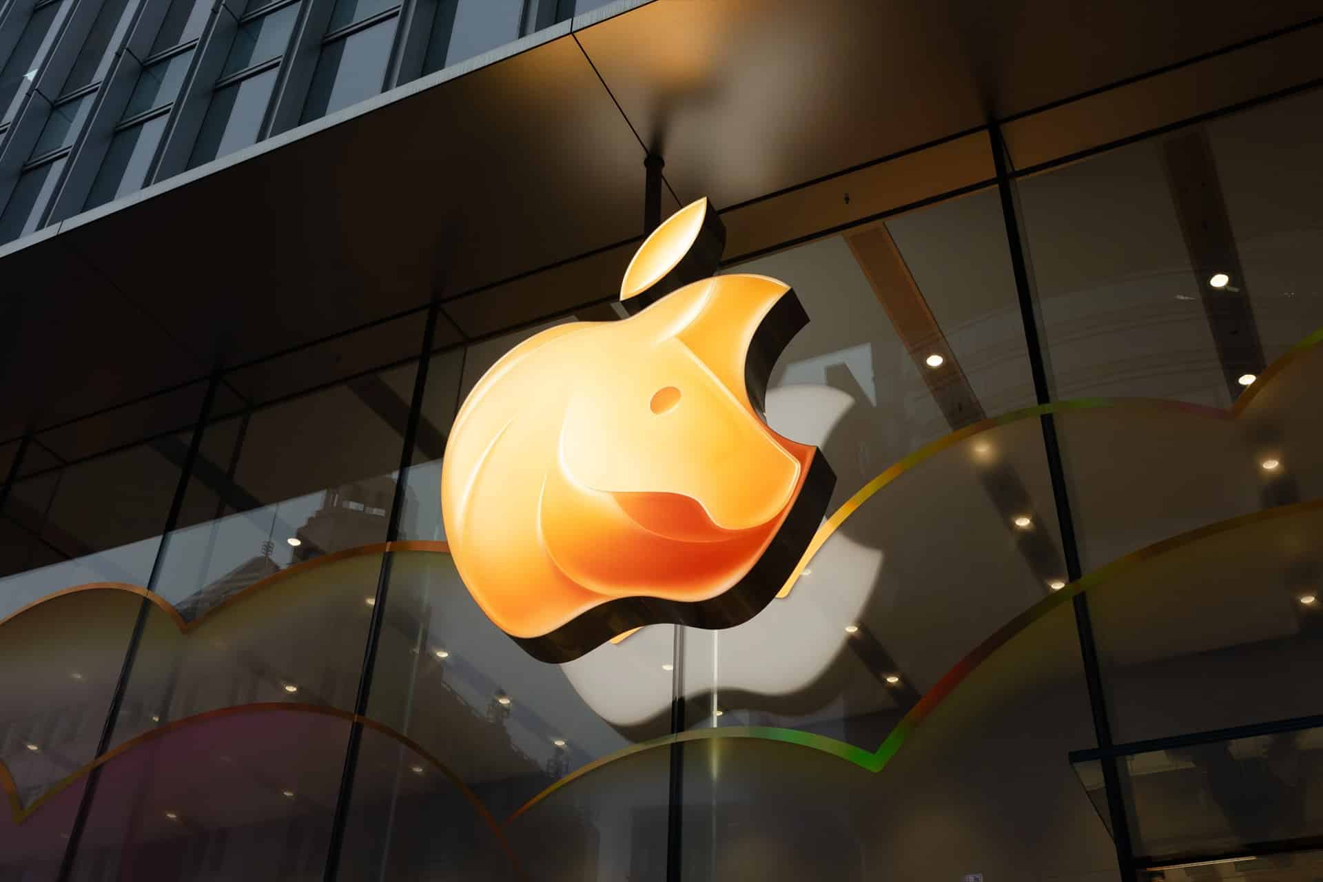 A large, illuminated Apple logo shines on the exterior glass facade of an Apple Store, with building reflections and colorful wavy lines celebrating Apple Lunar New Year along the bottom.
