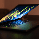 A partially closed laptop sits on a dark desk, its keyboard illuminated and screen displaying colorful, abstract lines—perhaps hinting at trends like apple stocks 2026. The background is blurred, focusing attention on the glowing device.