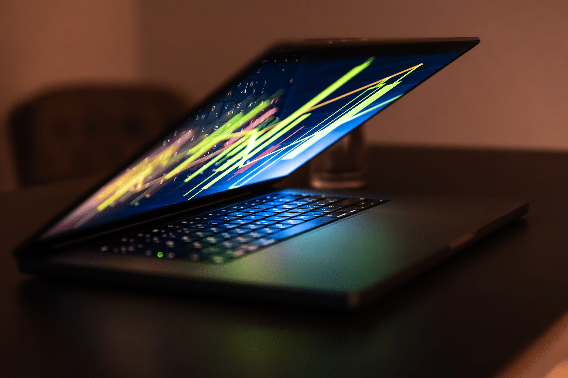 A partially closed laptop sits on a dark desk, its keyboard illuminated and screen displaying colorful, abstract lines—perhaps hinting at trends like apple stocks 2026. The background is blurred, focusing attention on the glowing device.