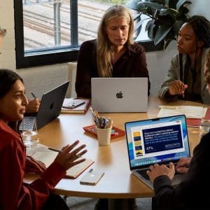 Five people sit around a round table in an office, discussing work with laptops, including a lightweight MacBook. One screen displays a colorful presentation slide as sunlight streams through the window.