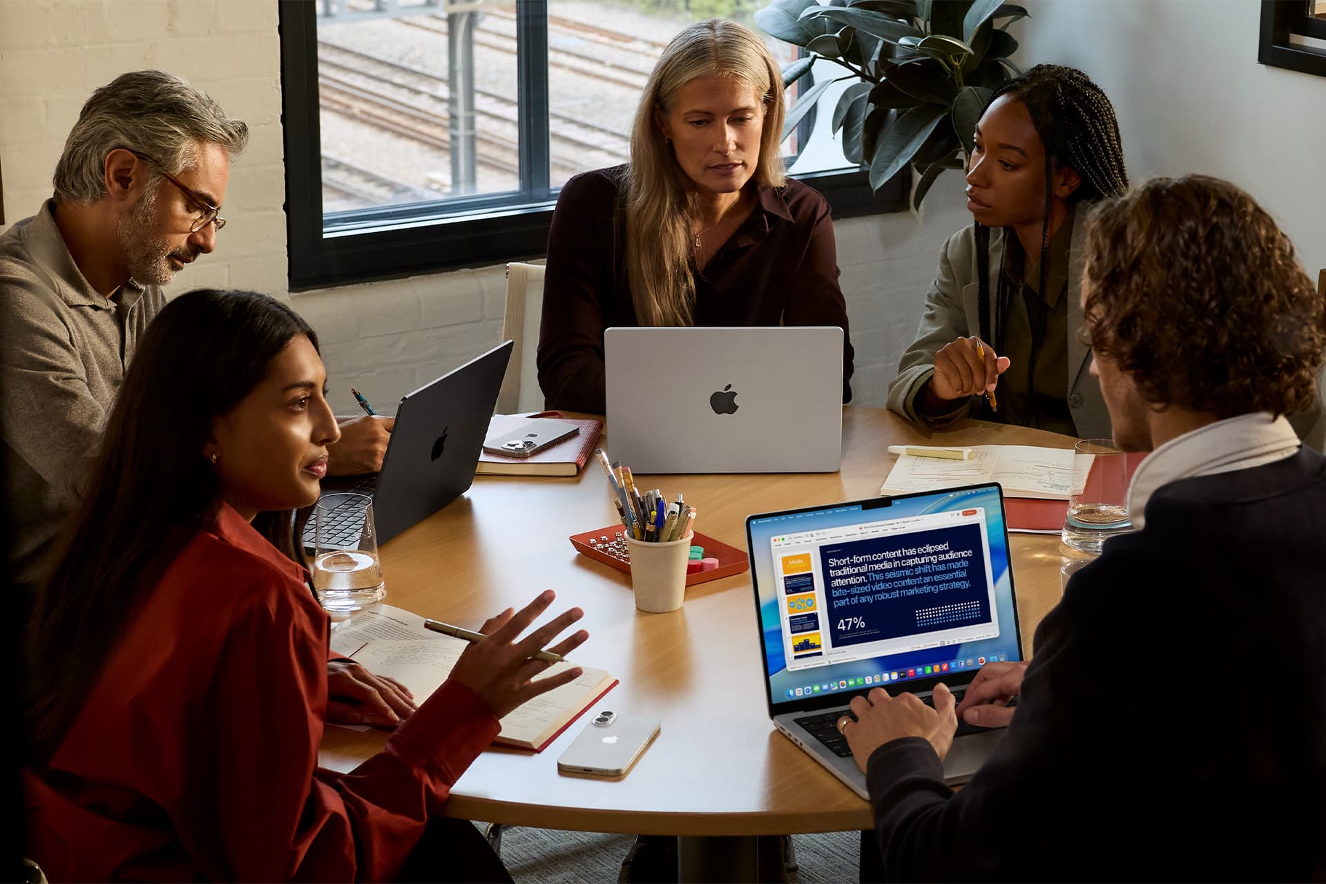 Five people sit around a round table in an office, discussing work with laptops, including a lightweight MacBook. One screen displays a colorful presentation slide as sunlight streams through the window.