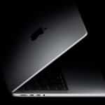 A partially open Apple MacBook laptop is shown in a dark setting, with the illuminated Apple logo visible on the lid and part of the keyboard, trackpad, and ports along the side in view.
