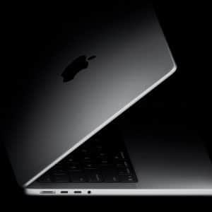 A partially open Apple MacBook laptop is shown in a dark setting, with the illuminated Apple logo visible on the lid and part of the keyboard, trackpad, and ports along the side in view.