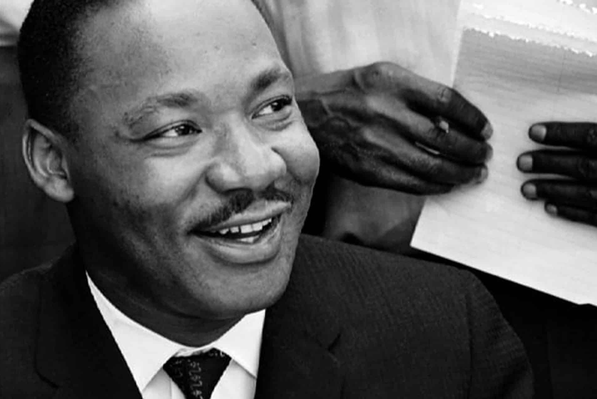 A Black man in a suit smiles while looking to the side, evoking the poised confidence of Martin Luther King. Behind him, another person's hands hold a notepad and pen. The image is in black and white.