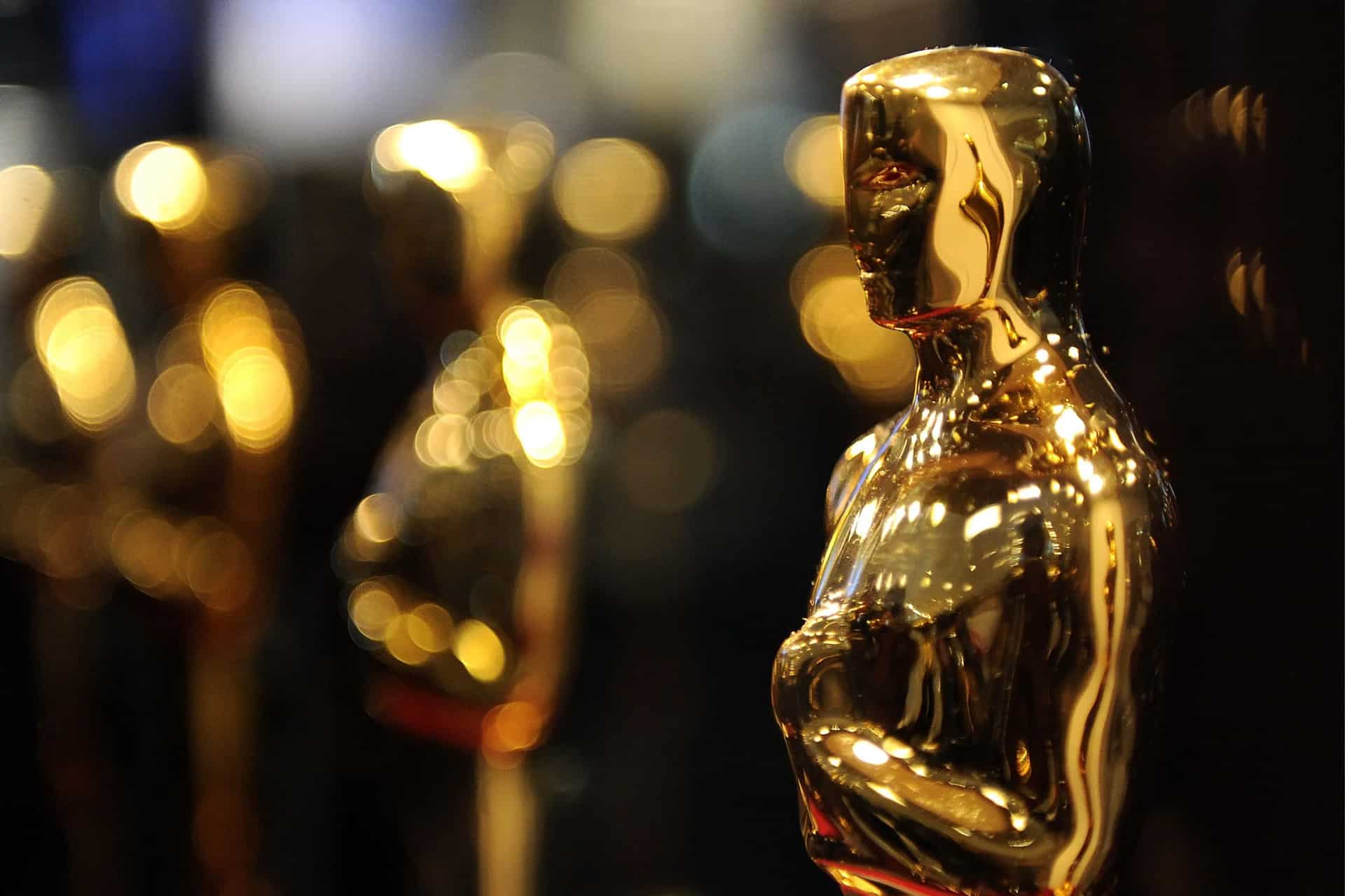 A close-up of a shiny gold Oscar statuette, with other similar statuettes blurred in the background against a backdrop of warm, out-of-focus lights—captured using Apple Creator Studio for stunning clarity. - Apple Academy Awards