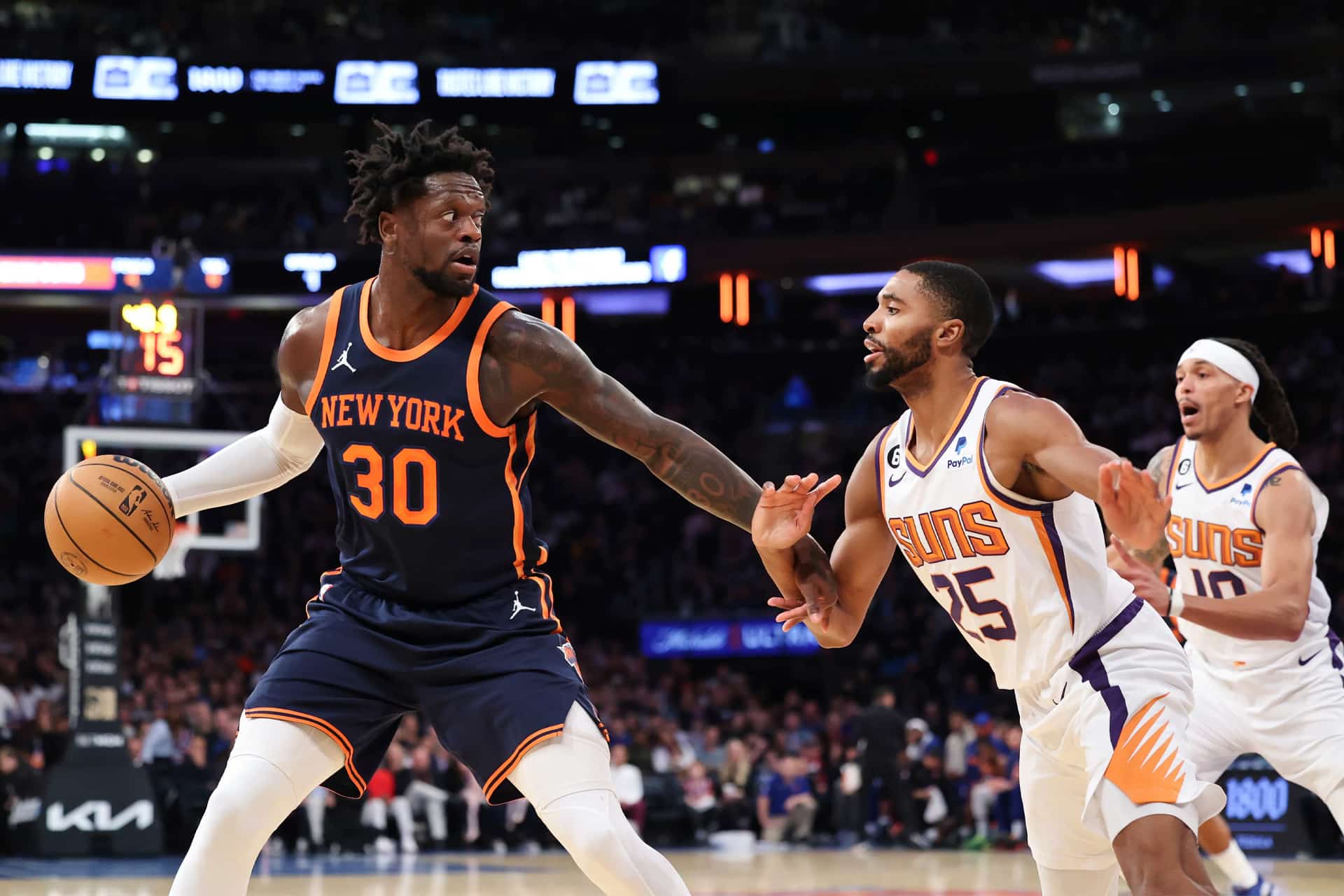 A New York Knicks player with the ball tries to drive past a Phoenix Suns defender, while another Suns player watches in the background during an NBA basketball game on Apple TV Game Night.