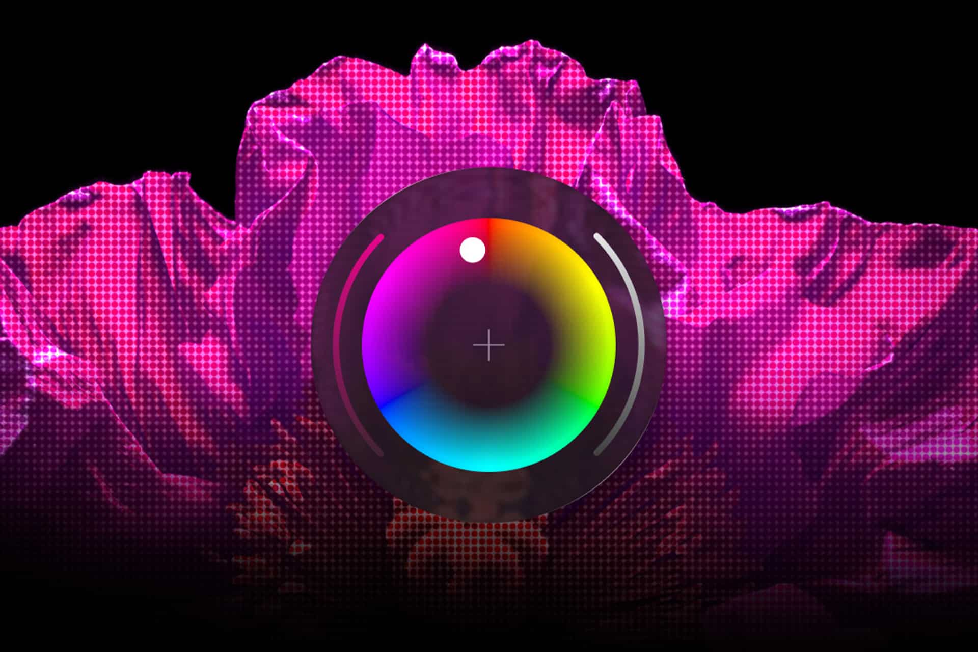 A large, bright pink flower with a pixelated texture is in the background. In the foreground, a circular color wheel resembling a camera lens—hinting at Apple vs Adobe's creative rivalry—is centered and overlaps the flower. The background is black.