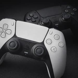 Two PlayStation 5 controllers, one white and one black, are placed side by side on a dark textured surface—ready for intense Apple TV Games sessions. The white controller leads in the foreground, with the black one slightly behind to the right.
