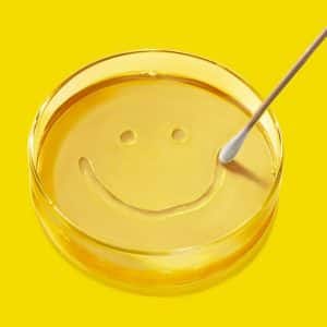Apple TV Pluribus Series | A cotton swab draws a smiley face in a petri dish filled with yellow liquid, set against a bright yellow background—perfect for fans of Season 2 or admirers of Carol's quirky Pluribus-inspired creations.