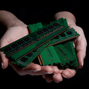 A pair of hands gently holds several green computer RAM sticks against a dark background, symbolizing the Memory Market Imbalance Driven by AI Demand.