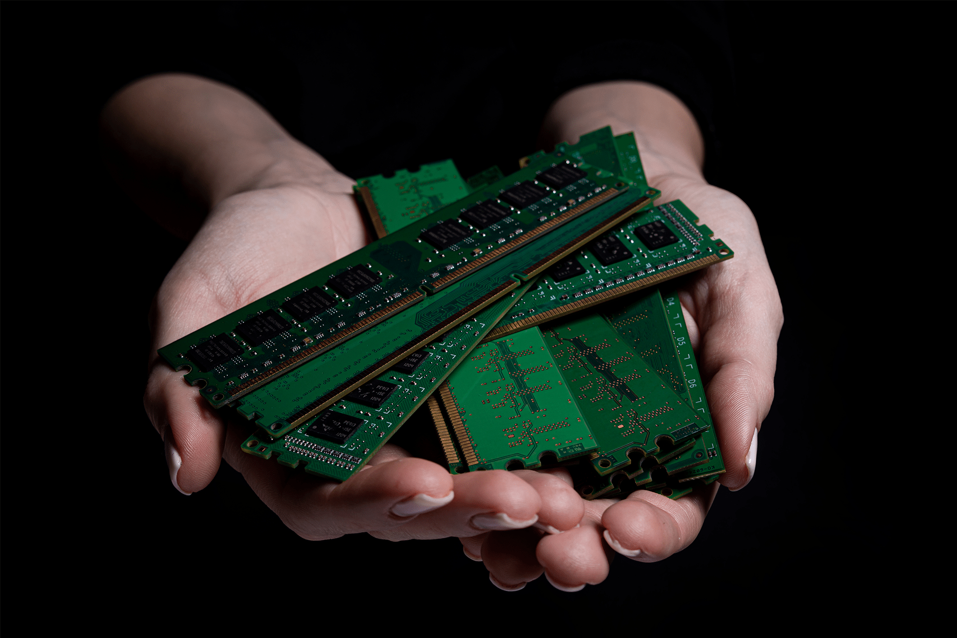 A pair of hands gently holds several green computer RAM sticks against a dark background, symbolizing the Memory Market Imbalance Driven by AI Demand.