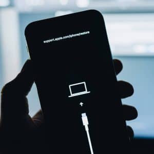 A hand holds an iPhone in iPhone Recovery Mode, displaying a restore screen with a cable pointing to a laptop icon and the URL support.apple.com/iphone/restore, indicating the device needs to be restored using a computer.