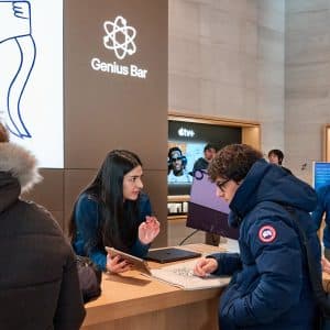 A woman at the Apple Sainte-Catherine Genius Bar assists a customer in a blue jacket, while others wait nearby; the Apple Montreal store features bright lighting and modern decor.