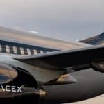 Starlink In Flight - Close-up of a sleek SpaceX jet featuring the Starlink In Flight logo on its shiny black fuselage and wing. The SpaceX branding stands out against the reflective surface, set beneath a cloudy sky.