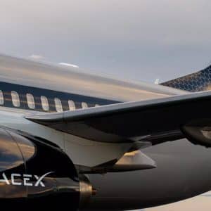 Starlink In Flight - Close-up of a sleek SpaceX jet featuring the Starlink In Flight logo on its shiny black fuselage and wing. The SpaceX branding stands out against the reflective surface, set beneath a cloudy sky.