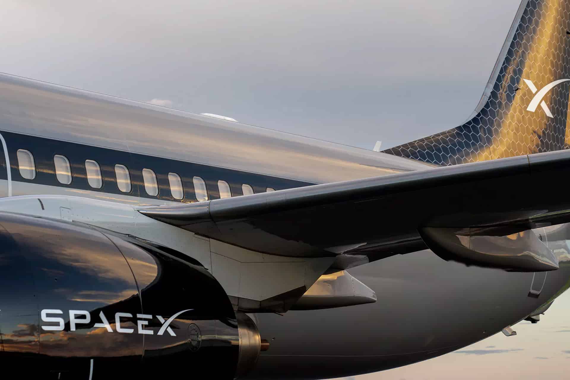 Starlink In Flight - Close-up of a sleek SpaceX jet featuring the Starlink In Flight logo on its shiny black fuselage and wing. The SpaceX branding stands out against the reflective surface, set beneath a cloudy sky.
