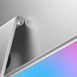 Close-up of the back of a silver Studio Display 2 monitor on a pink and blue gradient background, showing its stand and ventilation grille. An Apple logo is visible in the lower right corner.