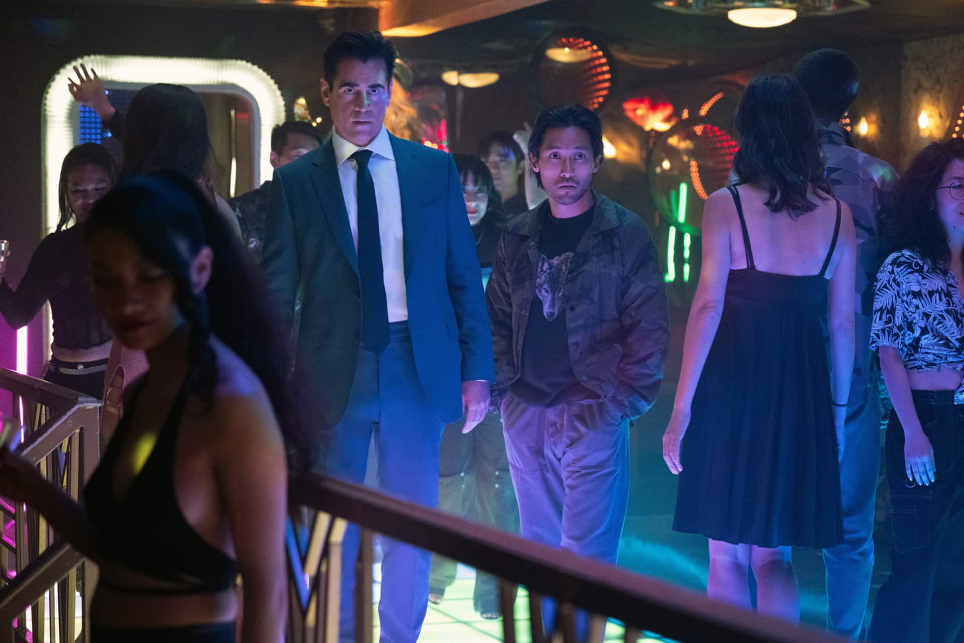 Two men, one in a blue suit and one in casual clothes, stand in a crowded, colorful nightclub surrounded by people dancing under neon lights—reminiscent of scenes from the Apple TV series Sugar Season 2.