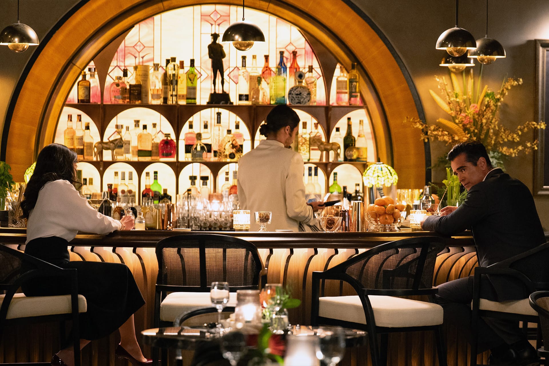 A woman and a man sit at a stylish bar with drinks, while a bartender stands behind the counter. The well-lit bar, featuring shelves of bottles and decorative lighting, feels like a scene from an Apple TV series such as Sugar Season 2.