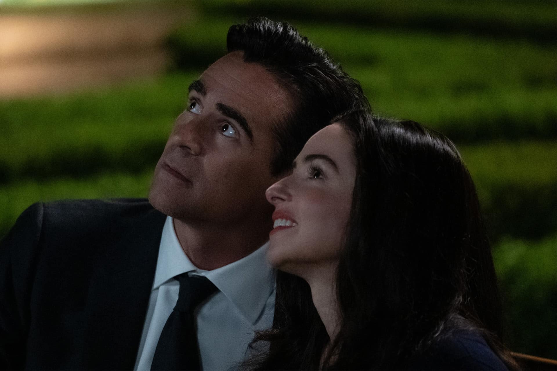 A man and a woman, both dressed formally, sit closely together outdoors at night, gazing upward and smiling—a scene reminiscent of heartfelt moments from Apple TV series like Sugar Season 2. The background is blurred with green grass and soft lighting.