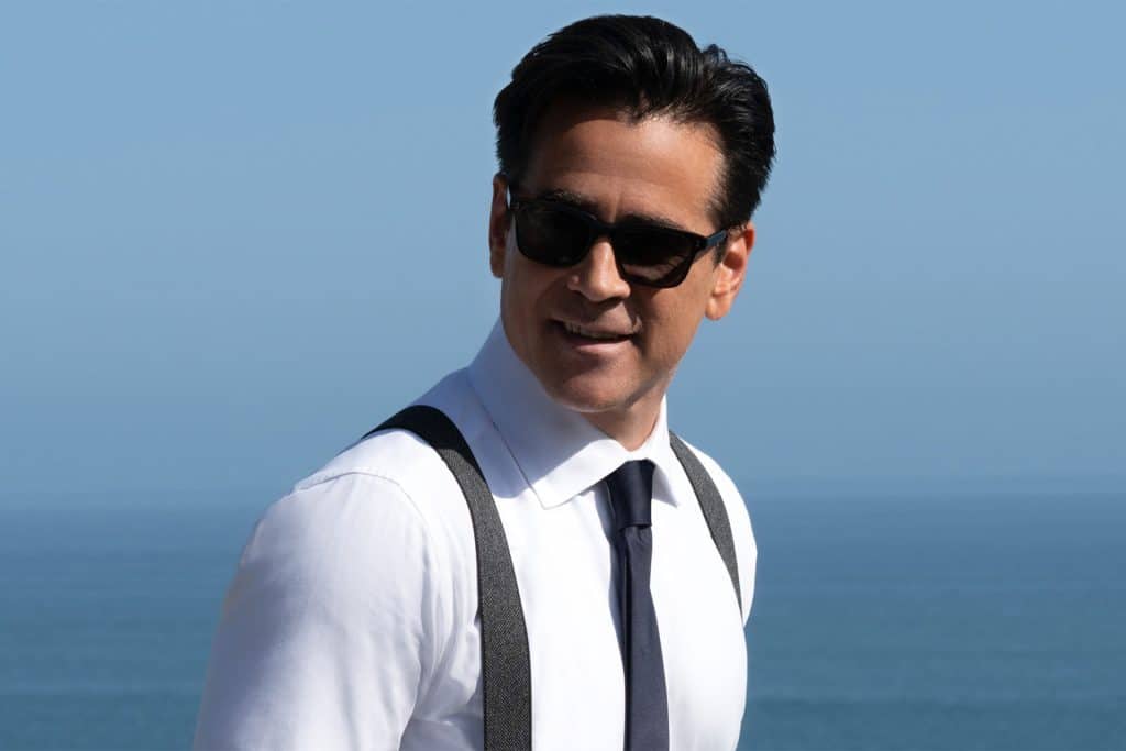 A man wearing sunglasses, a white dress shirt, black suspenders, and a dark tie stands outdoors with the ocean and blue sky in the background, reminiscent of characters from an Apple TV series like Sugar Season 2.