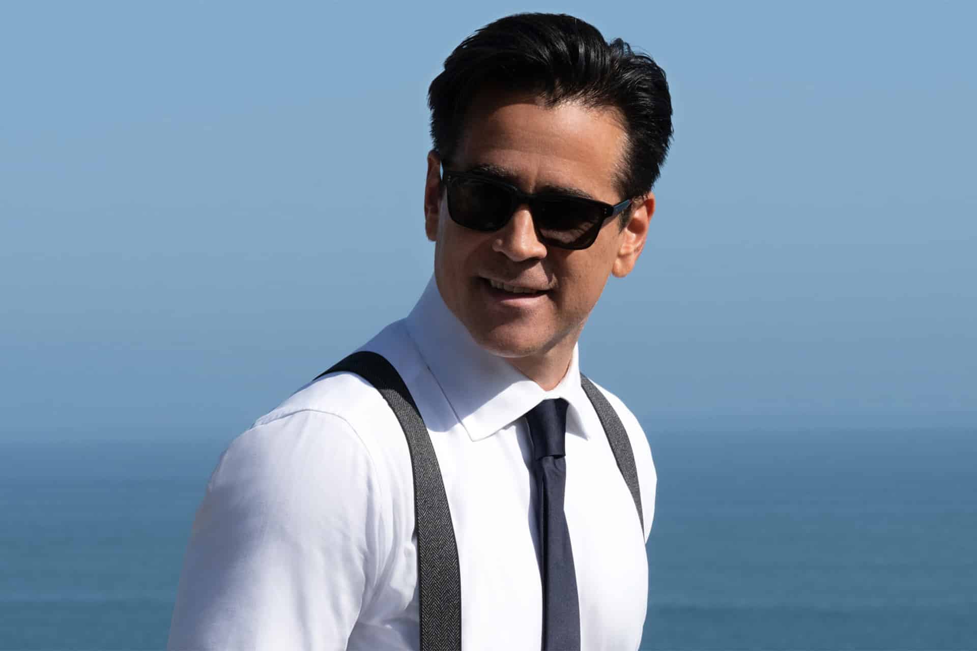 A man wearing sunglasses, a white dress shirt, black suspenders, and a dark tie stands outdoors with the ocean and blue sky in the background, reminiscent of characters from an Apple TV series like Sugar Season 2.