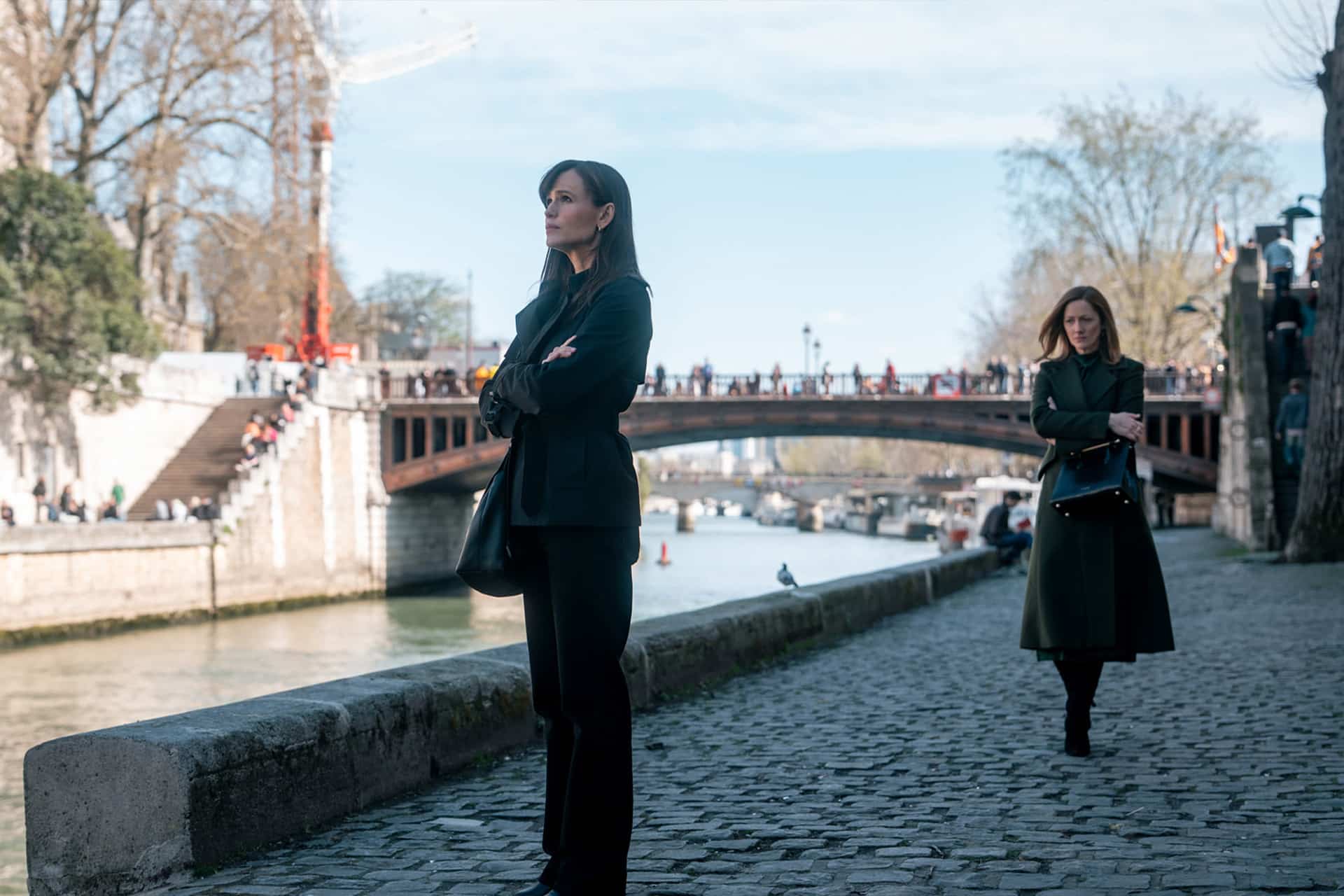 Two women walk along a cobblestone path by a river, one standing with arms crossed as the other approaches from behind. A bridge and people are visible in the background under a clear sky, evoking a scene from Last Thing Season 2.