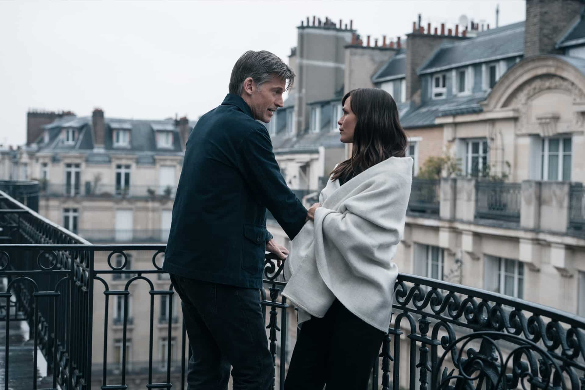 A man and a woman stand on a balcony, talking quietly as the woman, wrapped in a light-colored blanket, gazes at him. Behind them, classic European-style apartments stretch beneath a gray sky—an evocative scene from Last Thing Season 2.