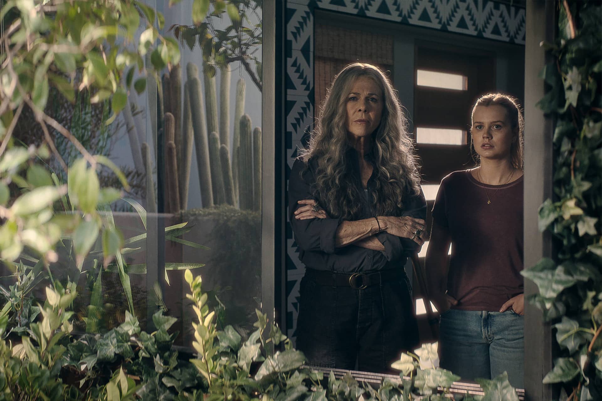Two women stand side by side inside a house, looking out through a glass window surrounded by green plants—a scene reminiscent of Last Thing Season 2. One has long gray hair and crossed arms; the other is younger with straight light hair, hands in her pockets.