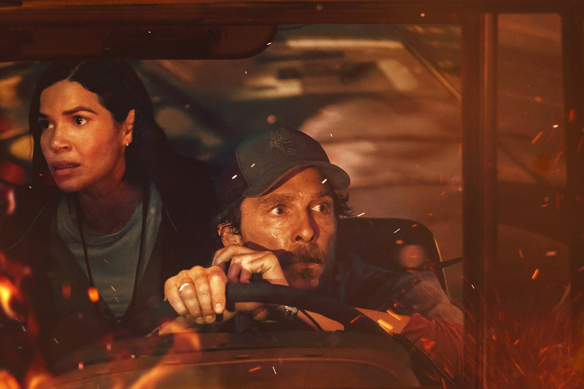 A woman and a man with intense, worried expressions sit in a vehicle, with the man gripping the steering wheel as flames and sparks—reminiscent of an Apple Creator Studio action scene—illuminate the windows outside.