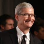 A man with short gray hair and glasses, resembling Tim Cook, smiles while standing indoors in a black suit. Two people are visible but out of focus behind him, hinting at an Apple 2026 "one more thing" announcement.