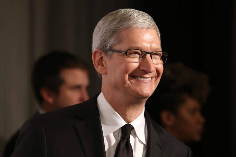 A man with short gray hair and glasses, resembling Tim Cook, smiles while standing indoors in a black suit. Two people are visible but out of focus behind him, hinting at an Apple 2026 "one more thing" announcement.