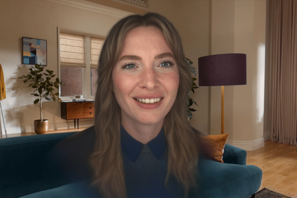 A woman with long brown hair and blue eyes smiles at the camera in a warmly lit living room, captured as a Vision Pro Persona, surrounded by plants, a lamp, artwork, and a window in the background.