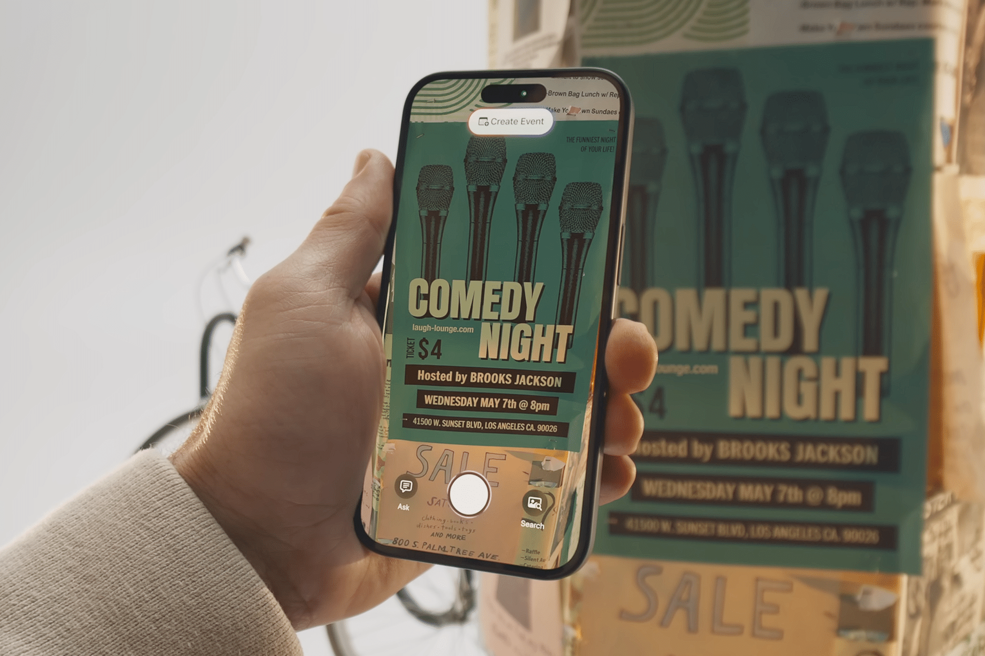 A hand holds a smartphone, capturing a photo of a comedy night poster. The phone screen displays the poster and camera interface, highlighting how visual intelligence works on iPhone to recognize details like microphones and event info.