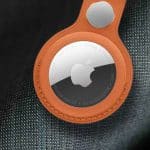 A close-up of an Apple AirTag in a brown leather keychain holder, attached to a textured gray fabric surface. The AirTag’s reflective surface and Apple logo are clearly visible. - New AirTag