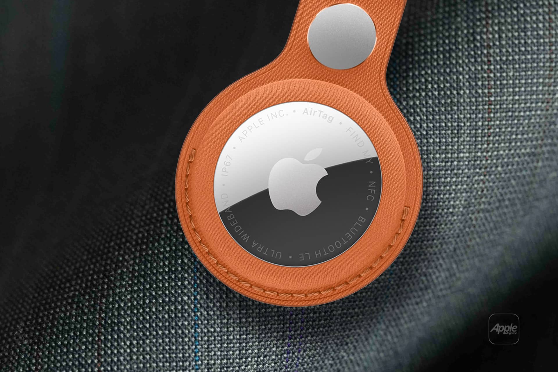 A close-up of an Apple AirTag in a brown leather keychain holder, attached to a textured gray fabric surface. The AirTag’s reflective surface and Apple logo are clearly visible. - New AirTag