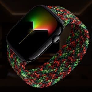 A close-up of an Apple Watch with a black case and a braided red, green, and black band, displaying a colorful gradient watch face, set against a dark, blurred background.