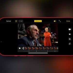 A smartphone screen displays a video editing app in iPhone Cinematic Mode, focusing on a man in a suit in the foreground and a woman in an orange outfit standing before bookshelves in the background.
