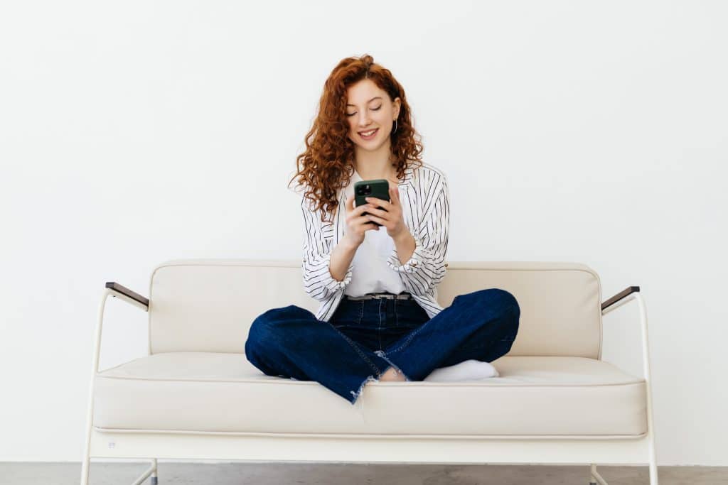 A young woman with curly red hair sits cross-legged on a light-colored sofa, smiling as she uses her iPhone as a remote control. She is wearing a striped shirt, white top, and blue jeans against a plain white background.