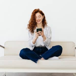 A young woman with curly red hair sits cross-legged on a light-colored sofa, smiling as she uses her iPhone as a remote control. She is wearing a striped shirt, white top, and blue jeans against a plain white background.